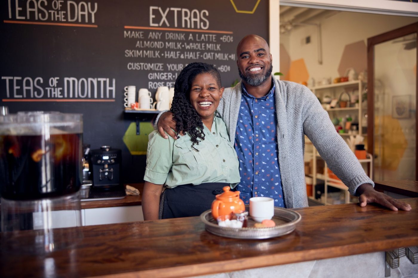 Male and female stood behind the counter of a cafe, menu part displayed behind them. they're smiling and the male has an arm over the shoulders of the female.