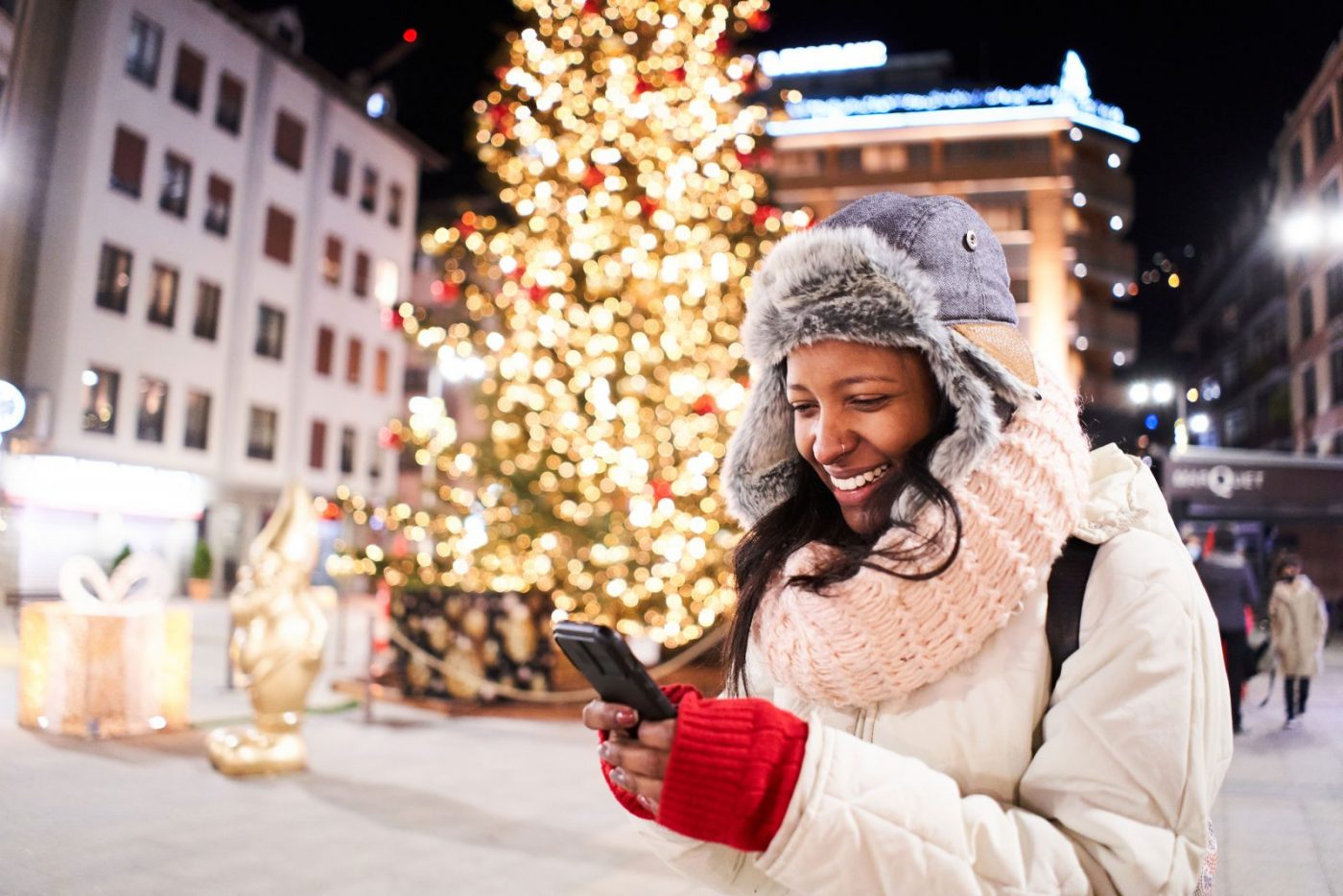 Woman wraped up from the cold, looking at her mobile phone in front of an outdoor Christmas tree with fairy lights.