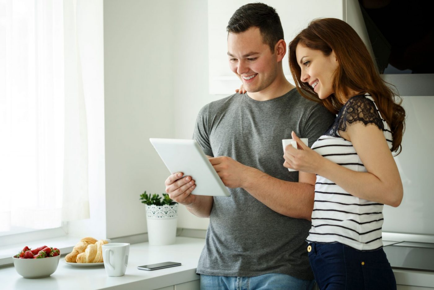 Male and female stood in a domestic kitchen setting, smiling as they drink from mugs and look at something on an ipad