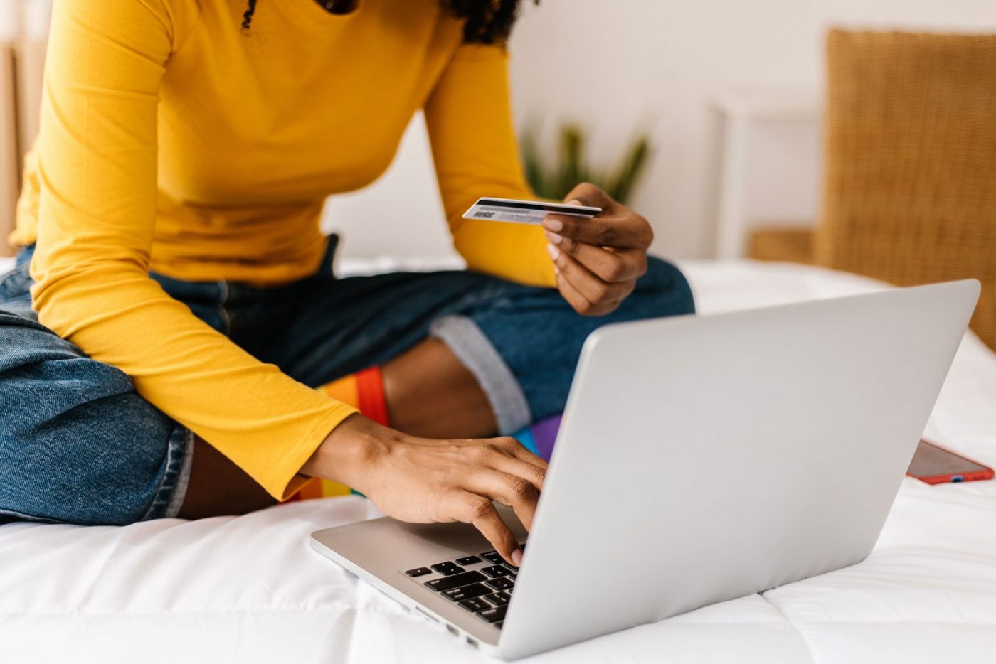 Image of a female from the shoulders down, wearing a yellow jumper and jeans, sat on a bed with a laptop open and a credit card in hand.