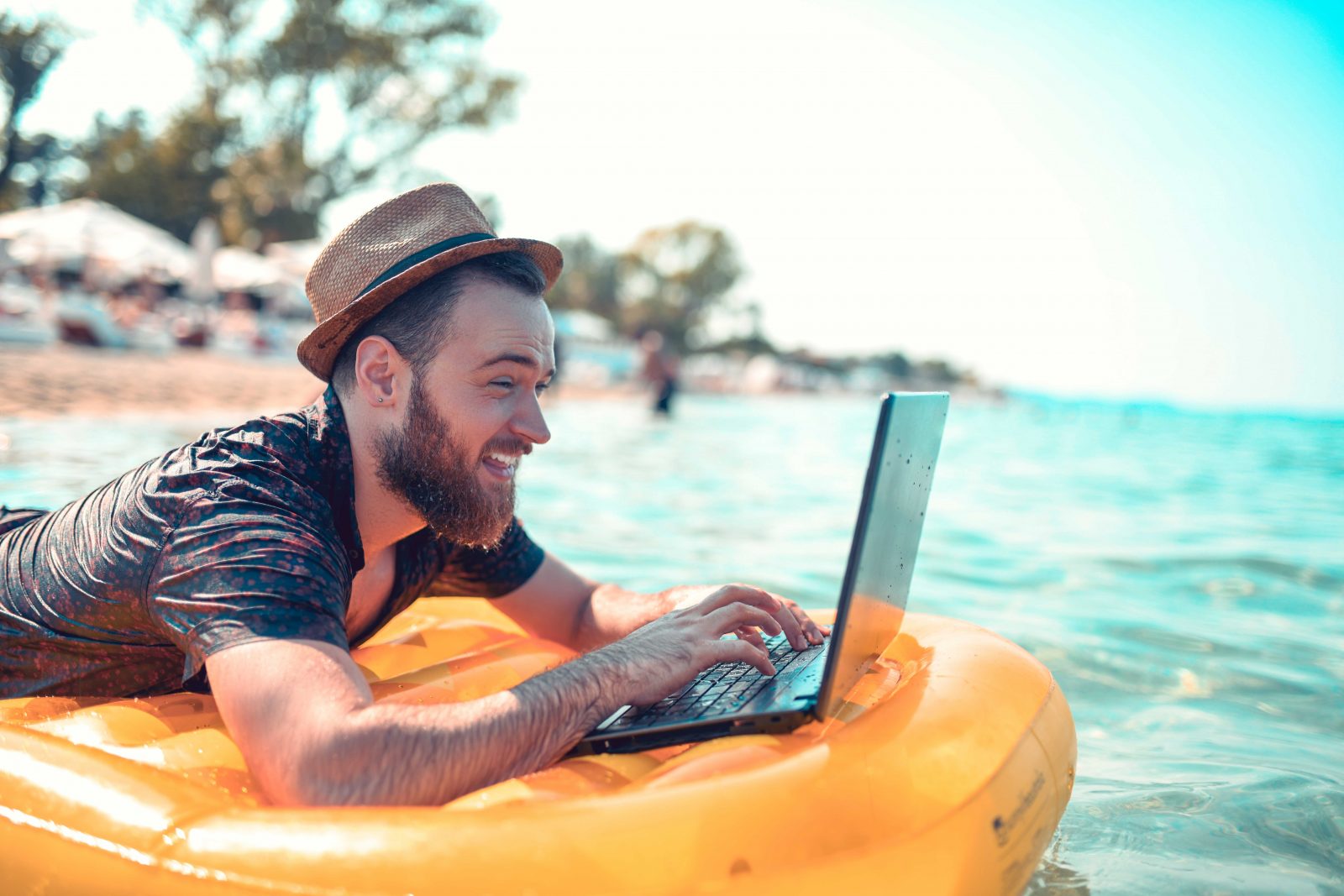 Image shows a man laid on an inflatable in the ocean whilst working on a laptop, the beach is visible behind him.