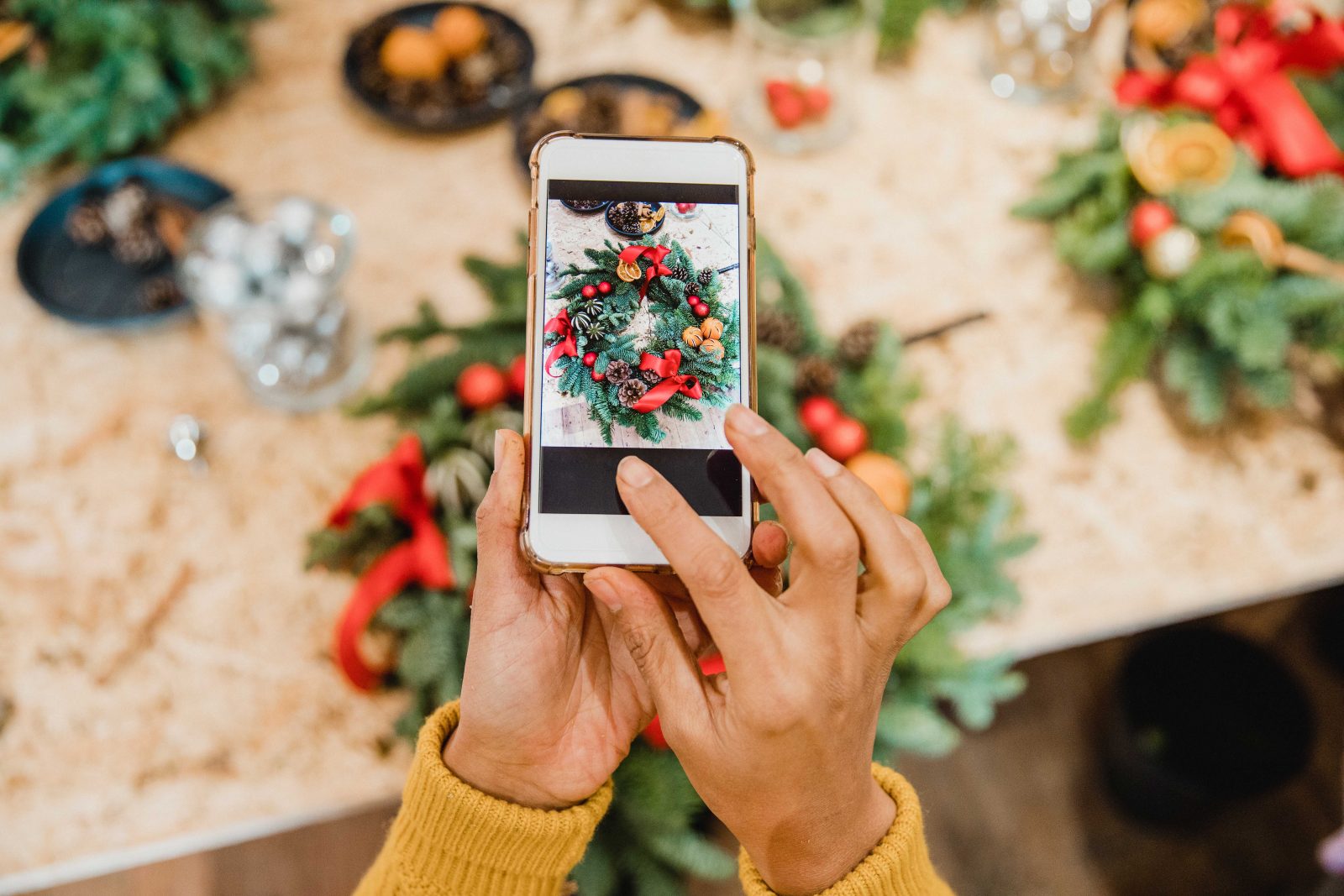 Image shows a pair of hands holding a mobile phone taking a photograph of a festive wreath