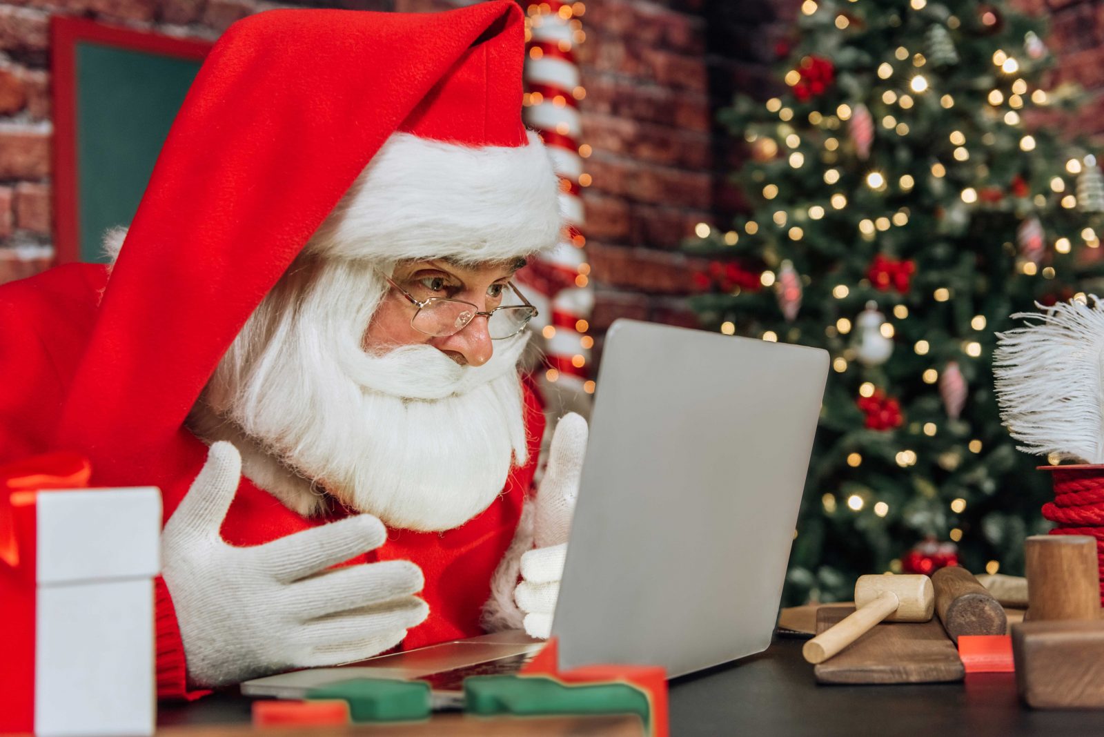 Image depicts a man dressed as Santa staring into a laptop screen, he looks excited and engaged by what he sees. The setting behind him is festive.
