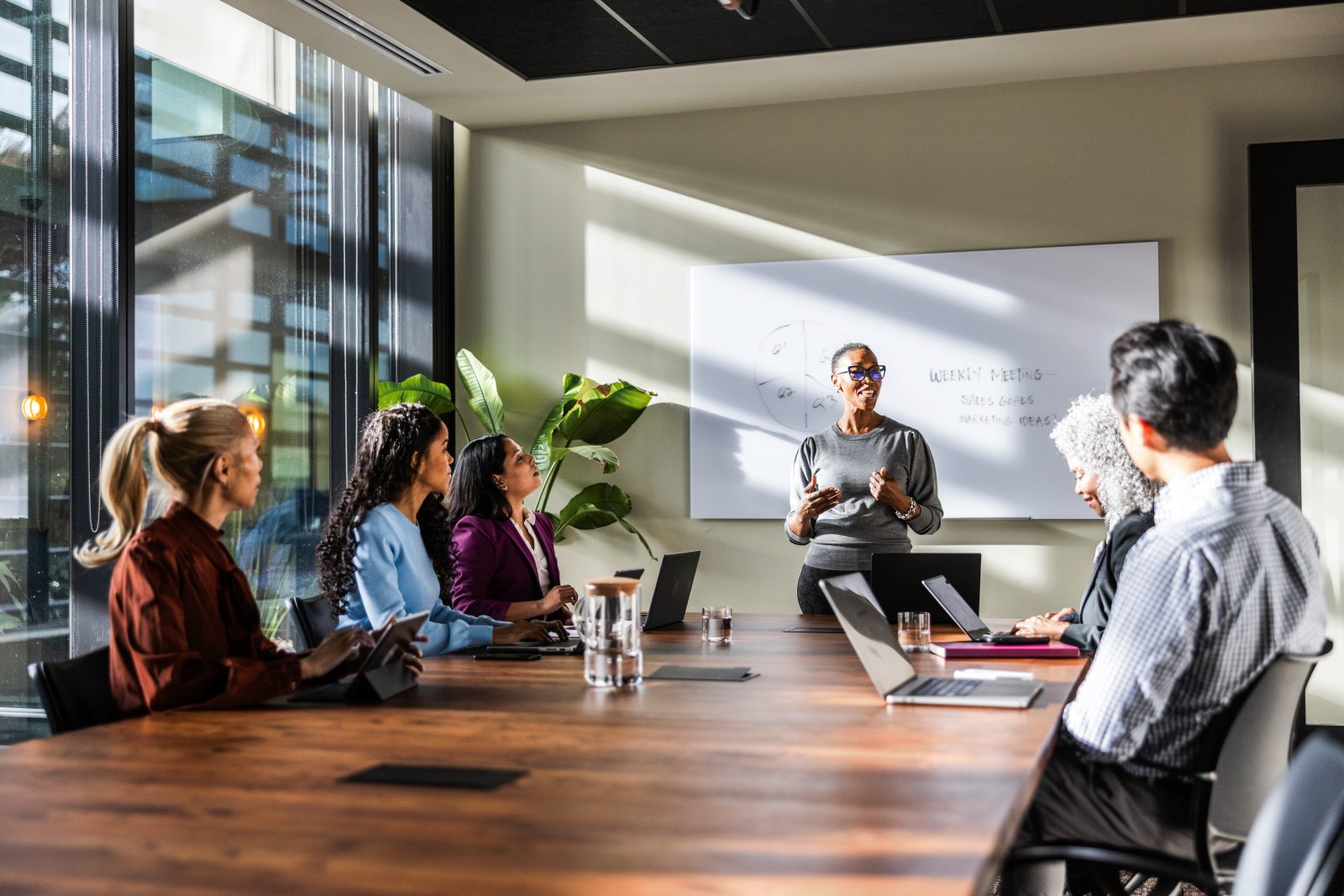 Image shows a meeting with a female stood at the head of a board table in front of a white board. Other people are sat around the table and looking at her as she addresses the room.