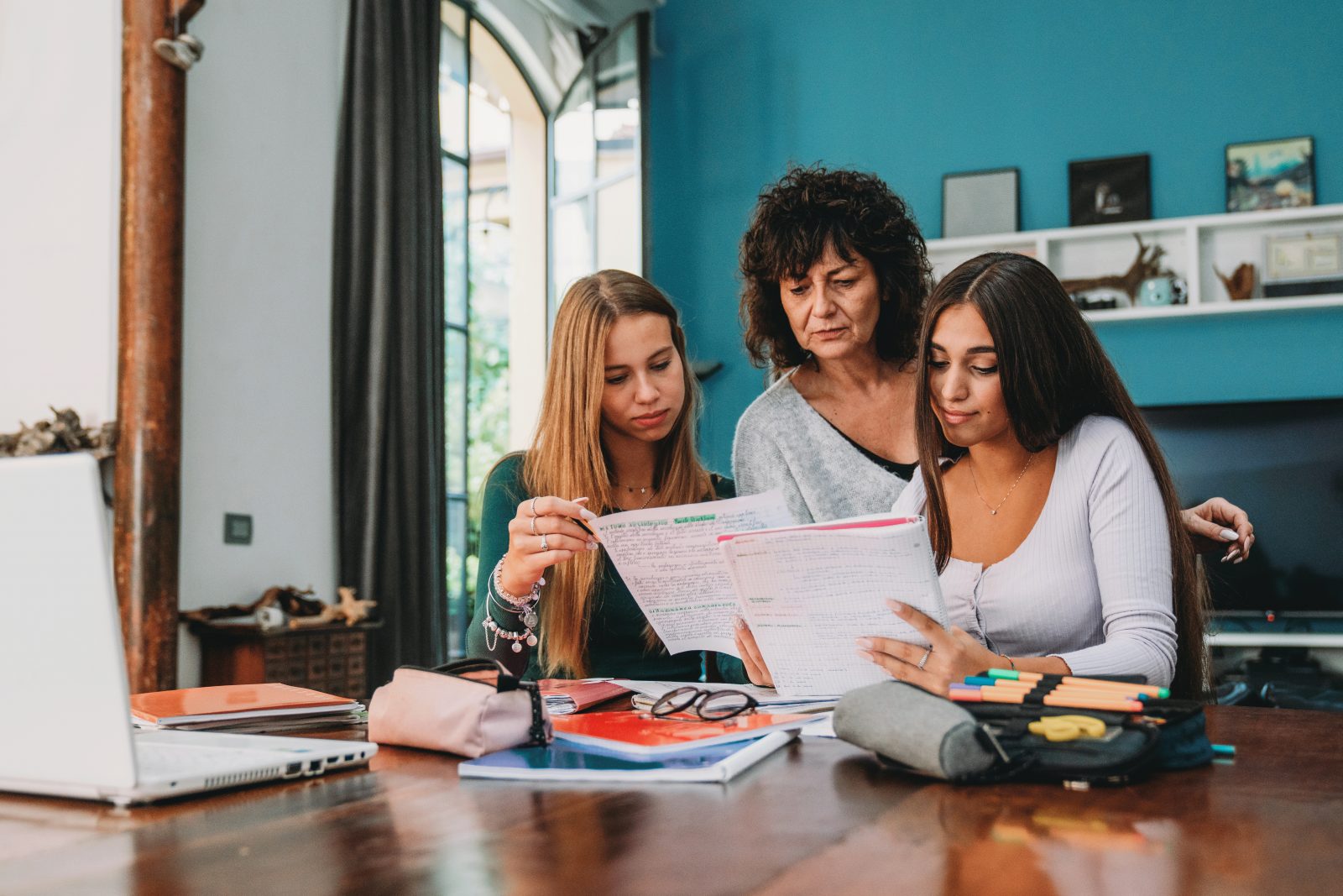 Image depicts an older female, standing over two teenage girls reading documents they are showing her. The younger girls are sat down at what looks like a dining table in a home setting. There are pens and papers on the table.