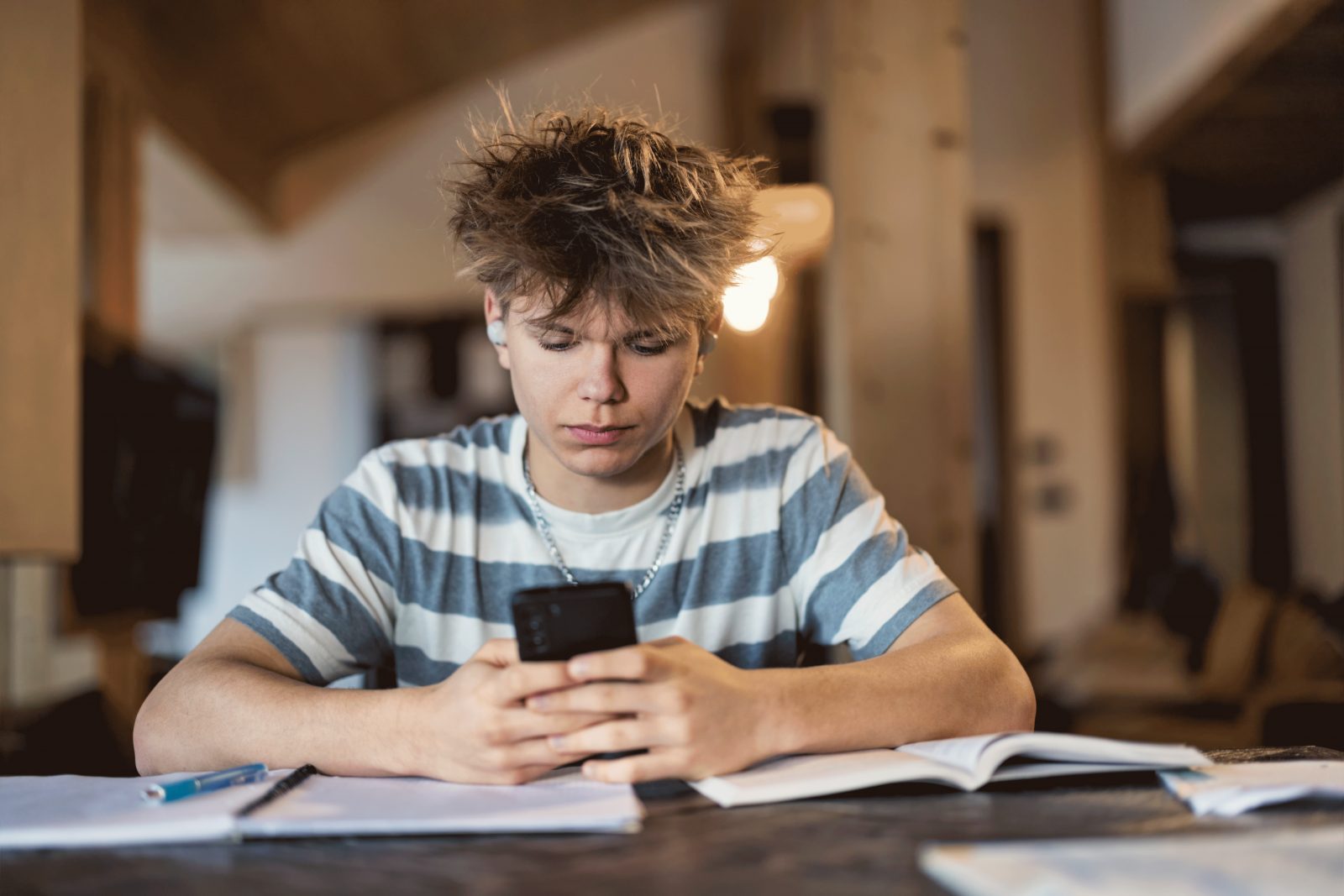 Teenage boy sitting at a wooden table, focused on his studies. The boy dressed in a gray and white striped shirt, is using his smartphone, listening to the music on his wireless in-ear headphones.