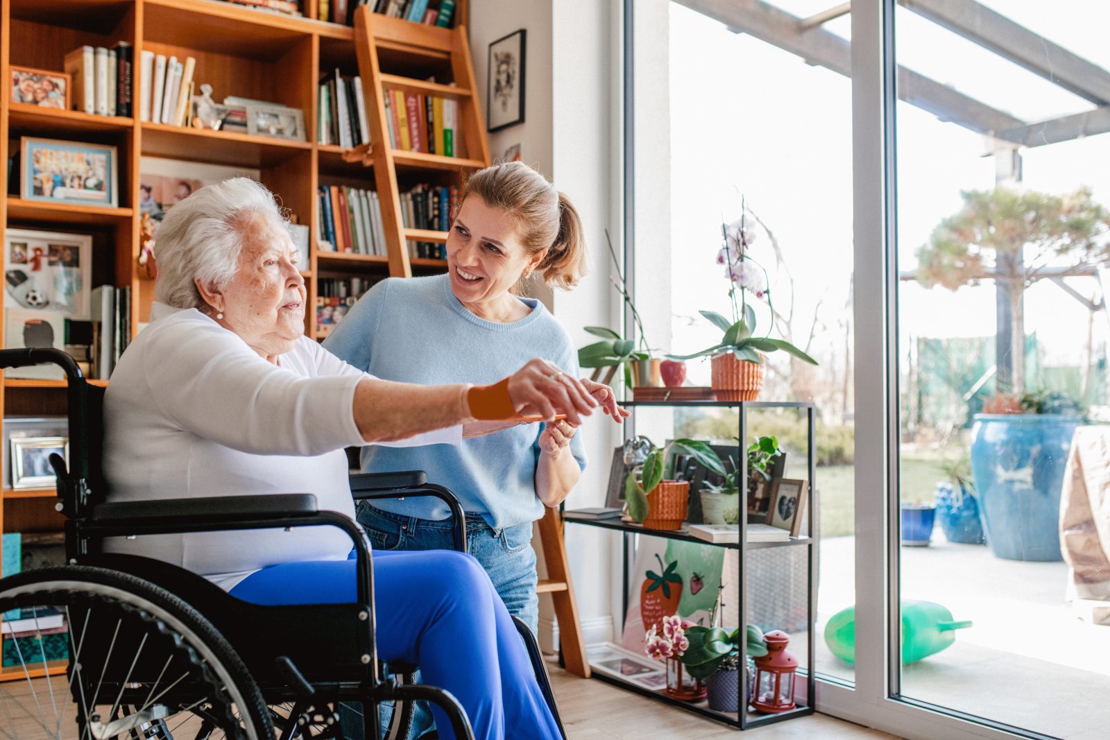 Adult daughter helping elderly mother in wheelchair with physiotherapy exercise at home.