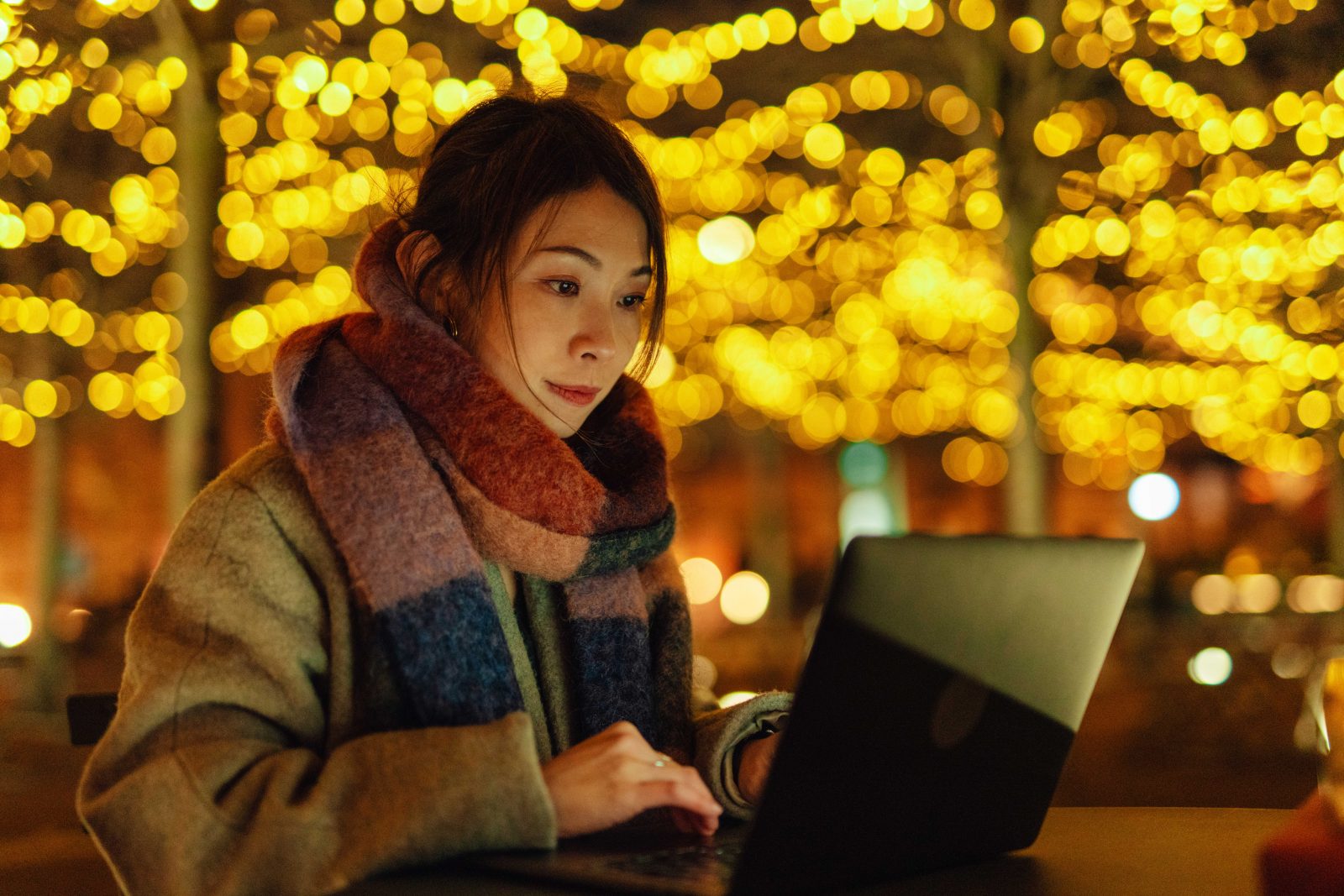 Young Asian woman working with laptop, sitting at sidewalk cafe, at night outdoors with bokeh street light in the background.